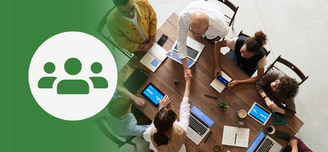 Overhead view of a diverse group of professionals seated around a large wooden table with laptops, notebooks, and coffee cups. Two people are shaking hands across the table. A green overlay on the left displays a white icon with three user figures—symbolizing teamwork or group collaboration.