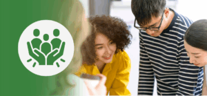 A group of diverse coworkers gathers around a table, engaged in discussion and problem-solving. One woman in a yellow shirt is smiling while leaning in. On the left, a green overlay features a white icon of three people enclosed by two protective hands—symbolizing team support, inclusion, or psychological safety.