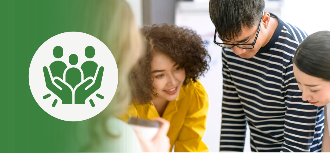 A group of diverse coworkers gathers around a table, engaged in discussion and problem-solving. One woman in a yellow shirt is smiling while leaning in. On the left, a green overlay features a white icon of three people enclosed by two protective hands—symbolizing team support, inclusion, or psychological safety.