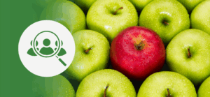 Close-up of apples with one red apple standing out among many green apples. On the left, a green overlay displays a white icon of a magnifying glass over a person’s head—symbolizing candidate search or identifying the right hire.